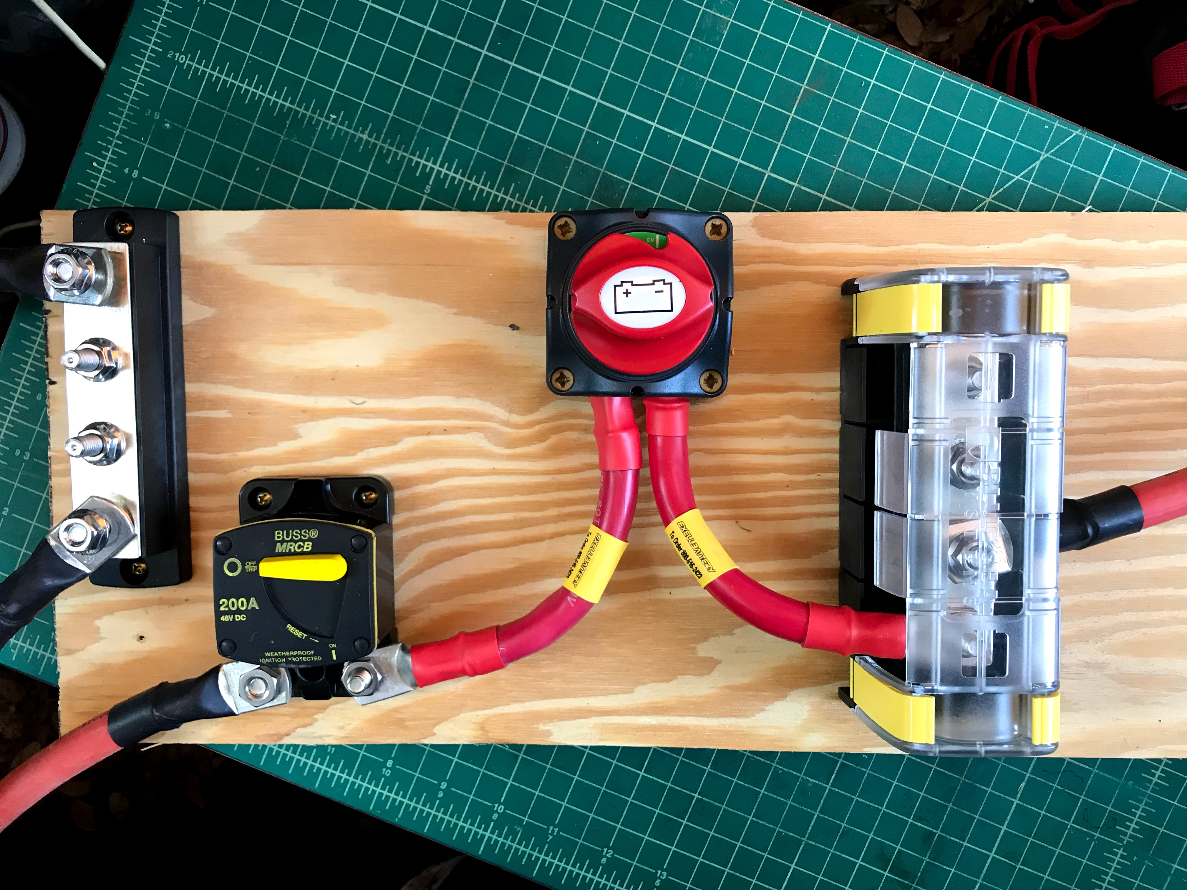 A  four post negative bus bar, 200 amp circuit breaker, battery cutoff switch, and 4 post positive bus bar with plastic cover are arranged left-to-right on and secured to a piece of plywood. Heavy 2/0 wires run between them. Red positive wires run to and from the breaker, cuttoff switch, and positive bus bar. Black negative wires run to the negative bus bar.
